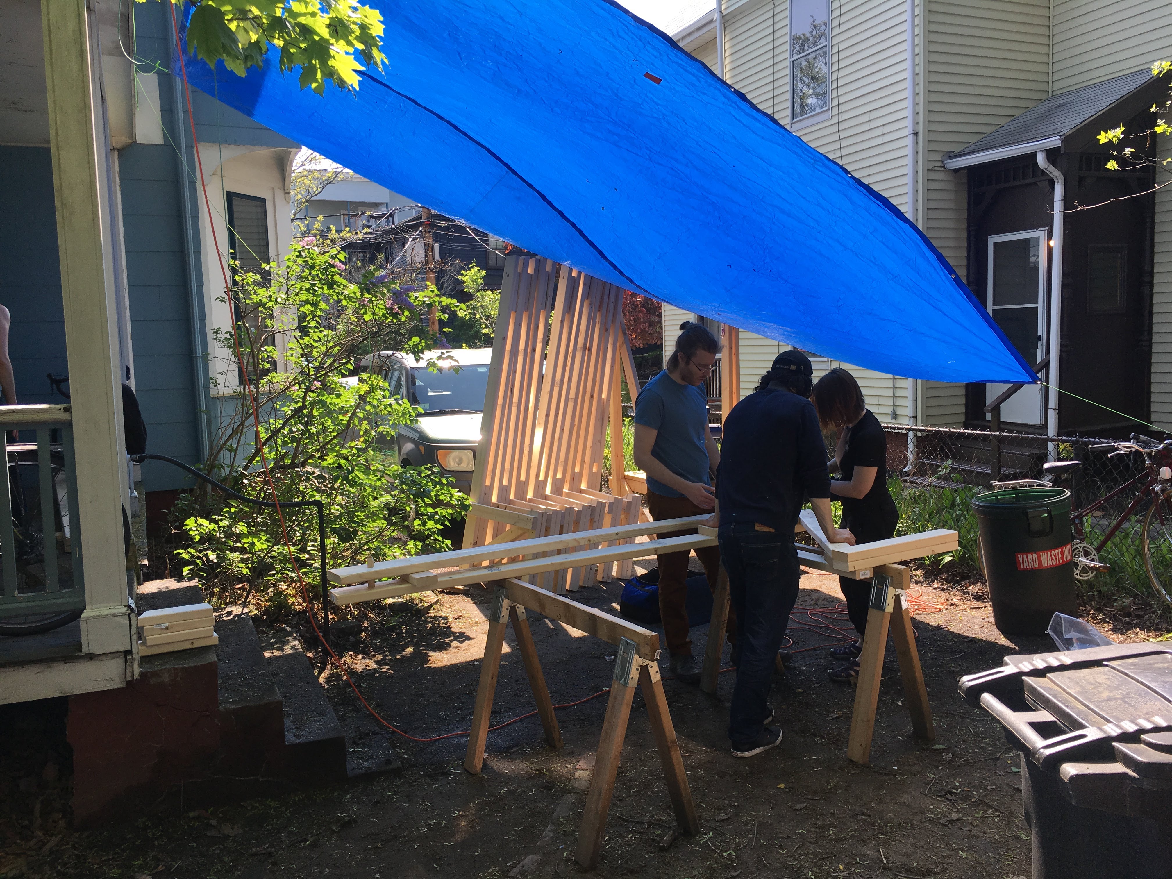 Crew members work wooden boards on sawhorses under a canopy
