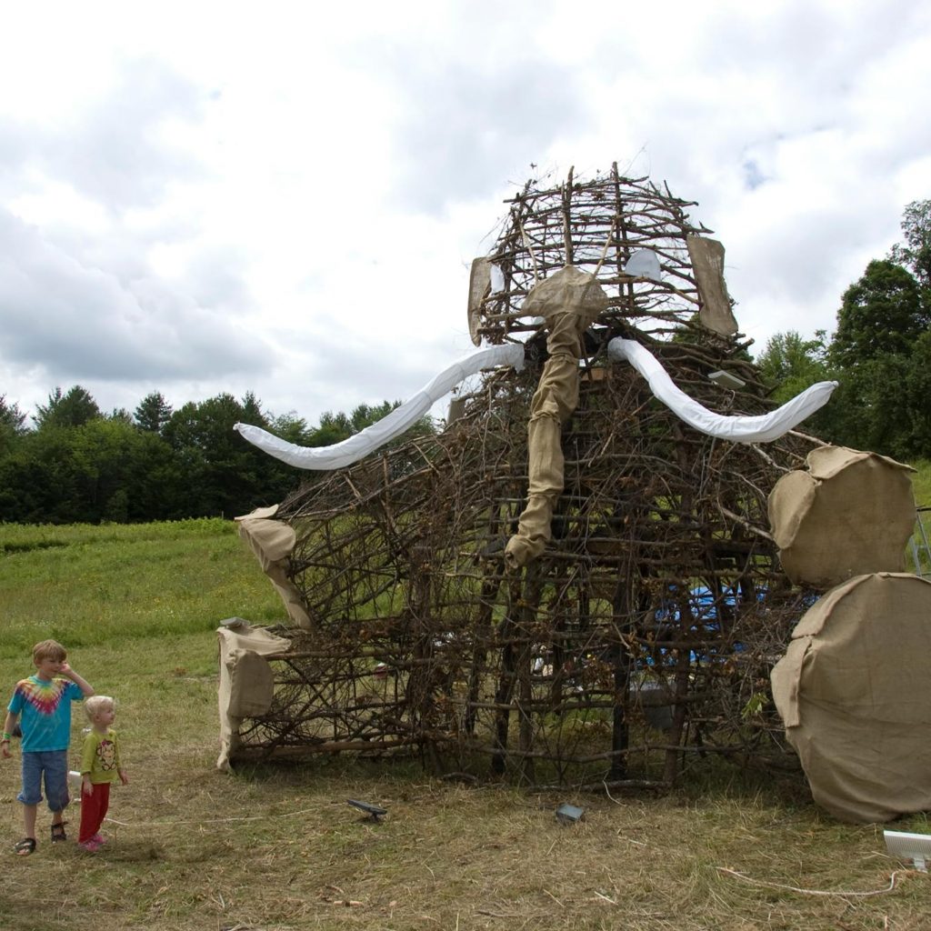 The 2011 bug during the day. A wooden effigy of a mammoth sitting on its rear. Its feet, trunk, ears and tusks are wrapped in cloth. An adult and two children admire it.